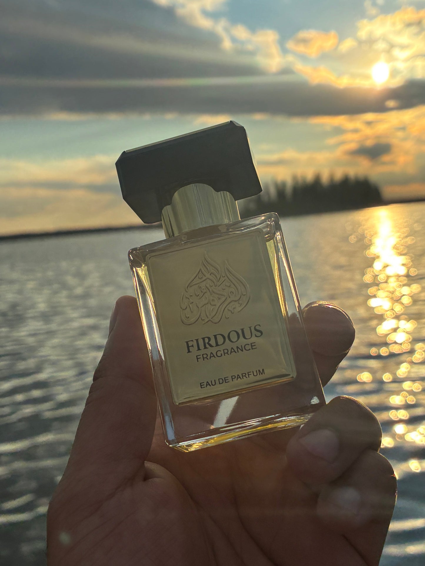 Hand holding a perfume bottle with a sunset and water background